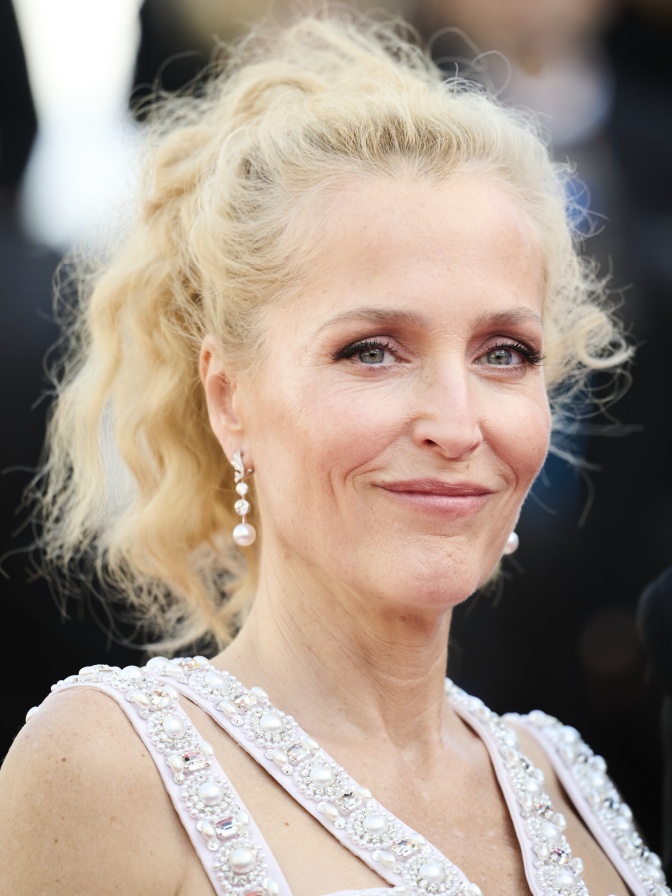 Gillian Anderson mit blonden, gewellten Haaren im Pferdeschwanz. Sie trägt ein festliches, glitzerndes Kleid und schaut direkt in die Kamera.