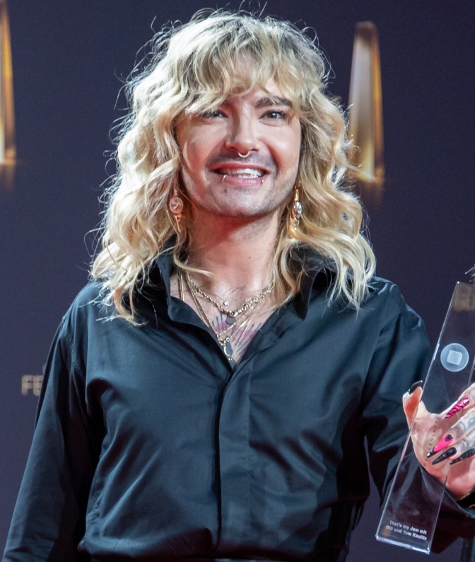 Bill Kaulitz mit langen, gewellten blonden Haaren in schwarzer Kleidung auf der Bühne, Er hält eine Preisstatue in der Hand und lächelt.