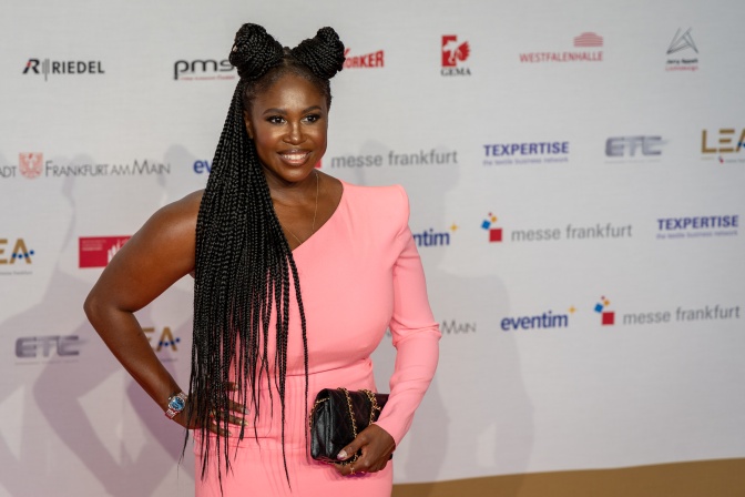 Motsi Mabuse mit zwei seitlichen Dutts. Sie trägt ein asymetrisches rosafarbenes Kleid und steht lächelnd vor einer Logowand.