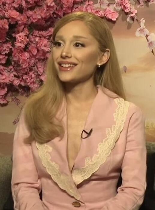 Ariana Grande in einem rosafarbenen Kostüm vor einer mit Blumen bewachsenen Wand.
