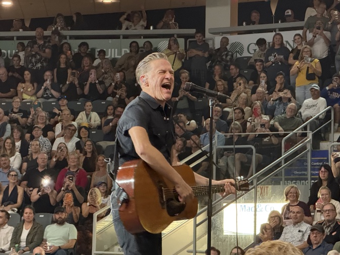Bryan Adams spielt Gitarre und singt. Er steht vor einem Mikrofon, rund um ihn herum Publikum.