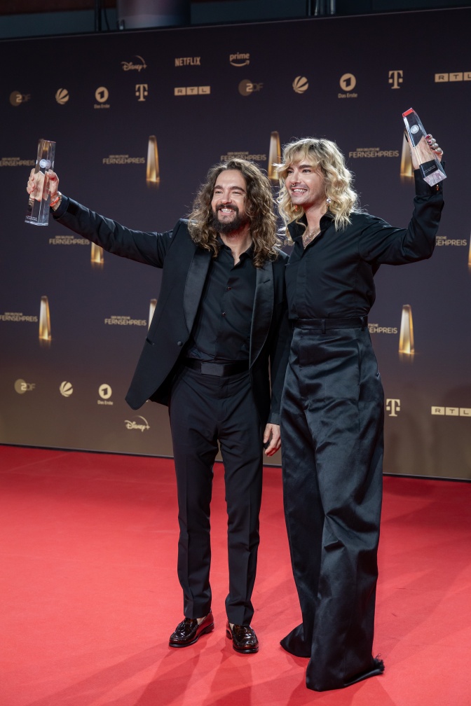 Bill und Tom Kaulitz stehen in schwarzen Anzügen auf dem roten Teppich und strecken eine Statue aus Plexiglas in die Höhe. Hinter ihnen eine Logowand.