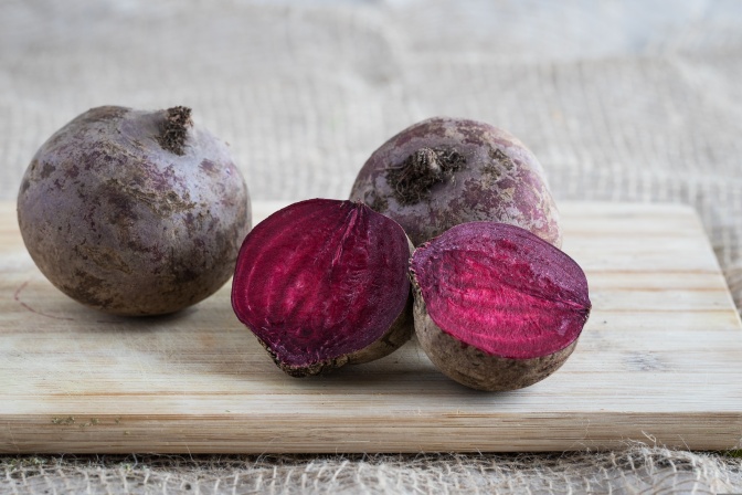 Auf einem Holzbrett liegen 3 Knollen Rote Beete, eine davon in der Mitte aufgeschnitten.