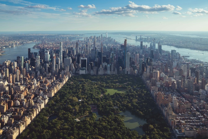 Der Central Park aus der Luft fotografiert. Eine Grünfläche mit Bäumen und Wiese, die Außenrum komplett von Hochhäusern umstanden ist.
