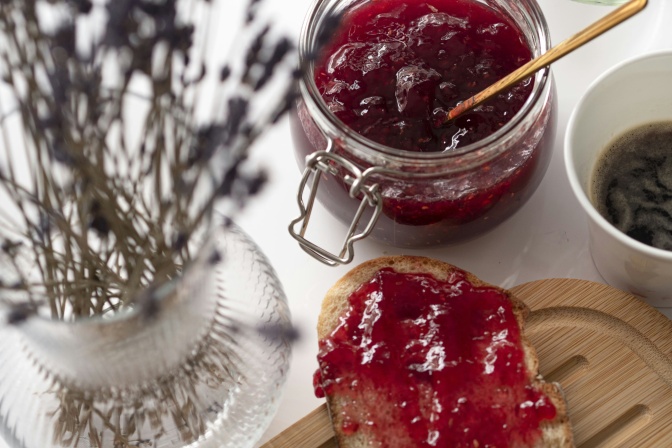 Ein Glas rote Marmelade mit einem Löffel darin, daneben liegt ein mit Marmelade bestrichenes Brot.