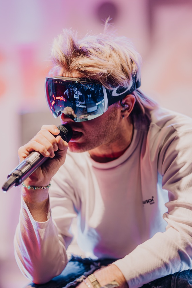 Ski Aggu it blondierten Haaren und spiegelnder Skibrille. Er hockt und singt in ein Mikrofon.