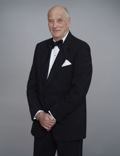 Ganzkörperfoto von König Harald in einem schwarzen Anzug mit Fliege. Er steht leicht seitlich zur Kamera und schaut ernst.