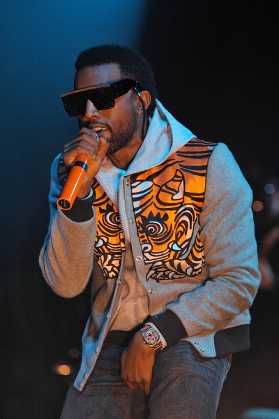 Kanye West auf der Bühne: Er trägt eine große, schwarze Sonnenbrille und eine bunte Jacke und singt in ein Mikrophon.