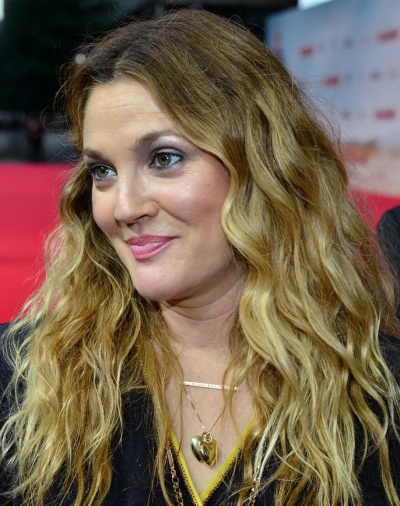 Drew Barrymore mit langen, gewellten, blonden Haaren und mit rosanem Lippenstift. Sie trägt eine goldene Kette mit 2 Herzen um den Hals.