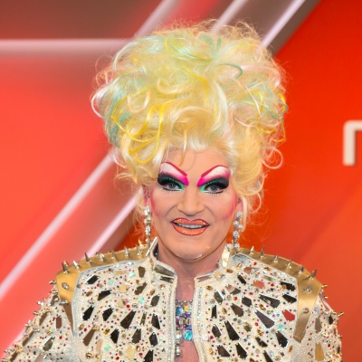 Olivia Jones in einer Jacke mit großen Glitzersteinen und mit einer hellgelben Perücke mit Turmfrisur. Sie ist stark geschminkt. Ihre Augen sind gelb, grün und rosa, ihre Lippen glitzern stark.