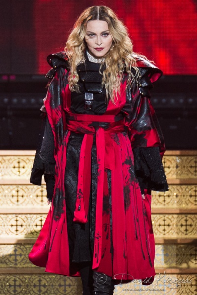 Madonna in einem rot-schwarzen Kleid auf der Bühne. Sie hat lange, gewellte, blonde Haare, die sie offen trägt. Sie geht eine Treppe herunter.