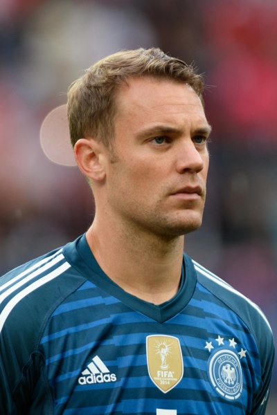Manuel Neuer mit einem Kurzhaarschnitt und in einem blauen Trikot mit drei weißen Streifen auf der Schulter und verschiedenen Markenlogos