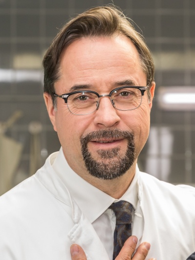 Jan Josef Liefers in weißem Kittel und mit Krawatte. Er trägt Brille und Vollbart.