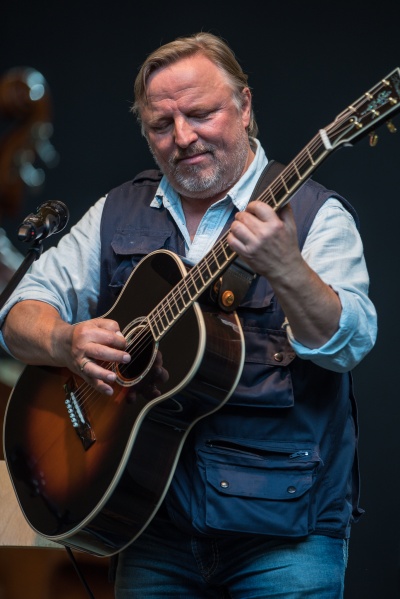 Axel Prahl trägt Hemd und Weste. Er spielt auf einer akustischen Gitarre.