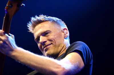 Bryan Adams in T-Shirt und mit kurzen, nach oben frisierten Haaren. Er steht vor einem Mikrophon.