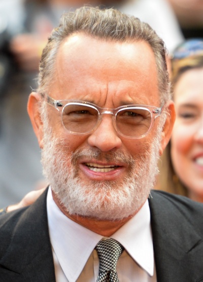 Tom Hanks mit grauen Haaren und weißem Bart. Er trägt Anzug und Krawatte und eine getönte Brille.
