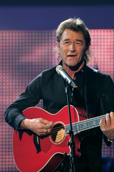 Peter Maffay mit kinnlangen Haaren. Er spielt Gitarre und steht vor einem Mikrophon.