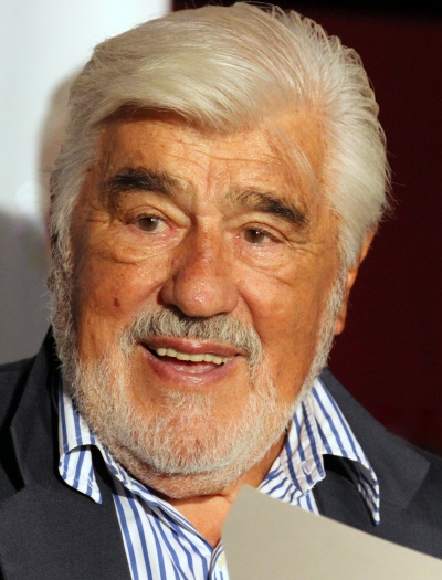 Mario Adorf mit weißen Haaren und weißem Bart. Er trägt Strickjacke und ein gestreiftes Hemd.