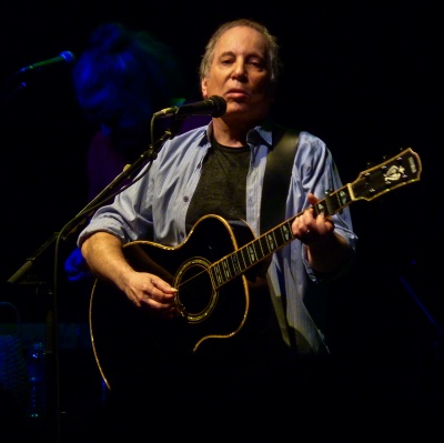 Paul Simon an einem Standmikro auf der Bühne. Er hat kurze Locken, spielt eine akustische Gitarre und singt.