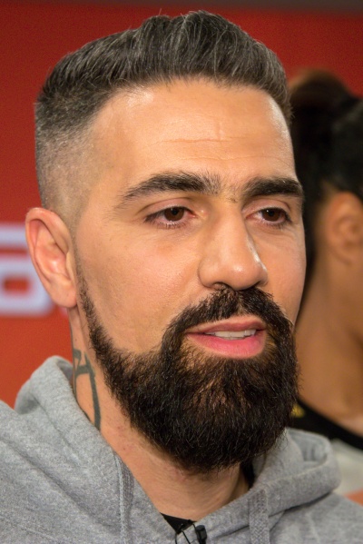 Bushido mit kurzen, an der Seite ausrasierten Haaren und mit Vollbart vor einer Logowand.