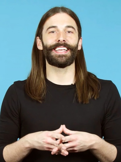 Jonathan Van Ness mit langen, glatten Haaren und Vollbart. Er hat die Hände vor dem Körper verschränkt und lächelt in die Kamera.
