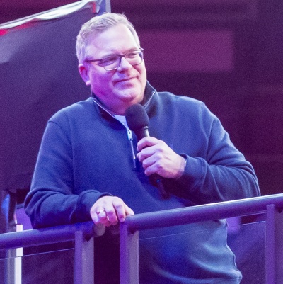 Elton lehnt in einem Wollpullover an einem Geländer und spricht in ein Mikrofon.