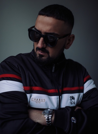 Haftbefehl mit einer dunklen Sonnenbrille und in einer Trainingsjacke. Er hat die Arme vor der Brust verschränkt und schaut zur Seite.